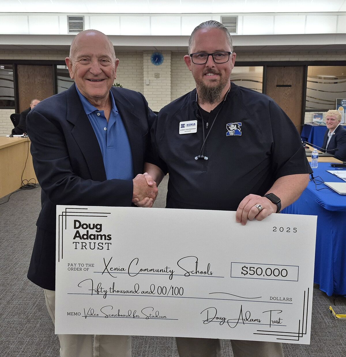Two men shake hands while holding a large check for Xenia Community Schools.