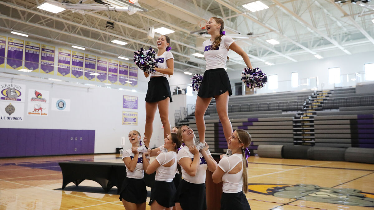 Cheerleaders perform a stunt in a gymnasium, with two girls standing on top of a pyramid.