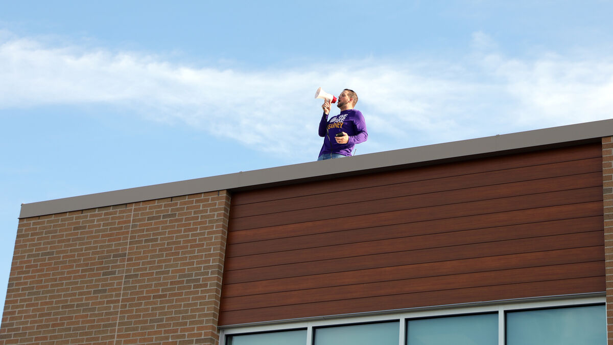 A man stands on a building roof, speaking into a megaphone under a blue sky.