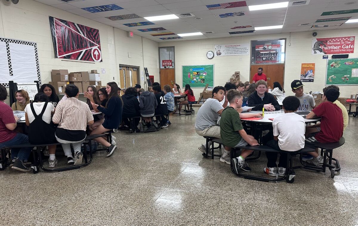 Students sit at round tables in a school cafeteria, engaged in conversation.