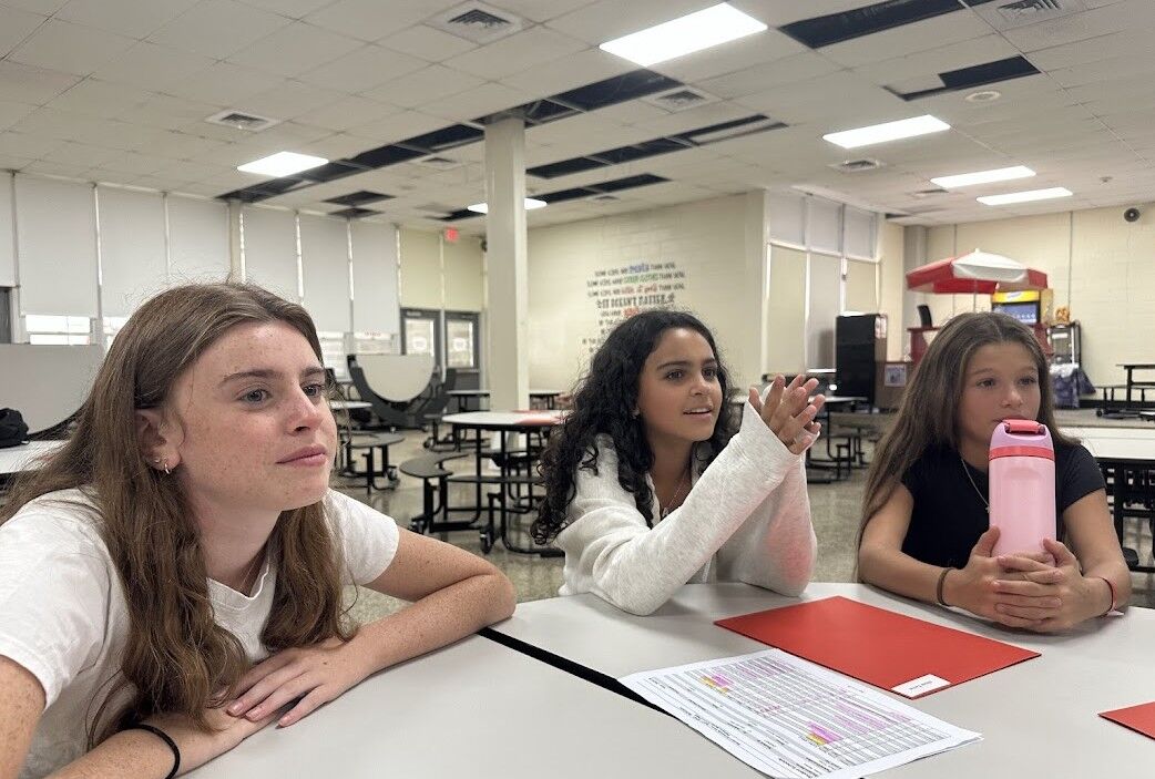 Three young people sit at a table in a school cafeteria, engaged in conversation.