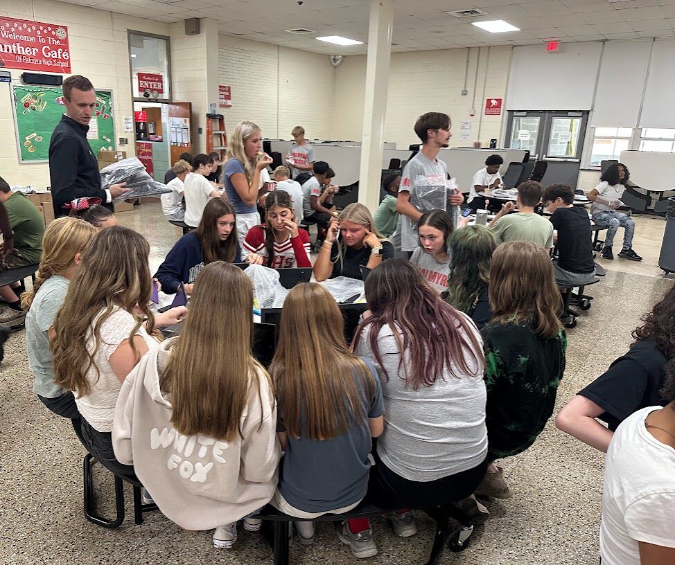 Students gather around tables in a school cafeteria, some looking at papers.