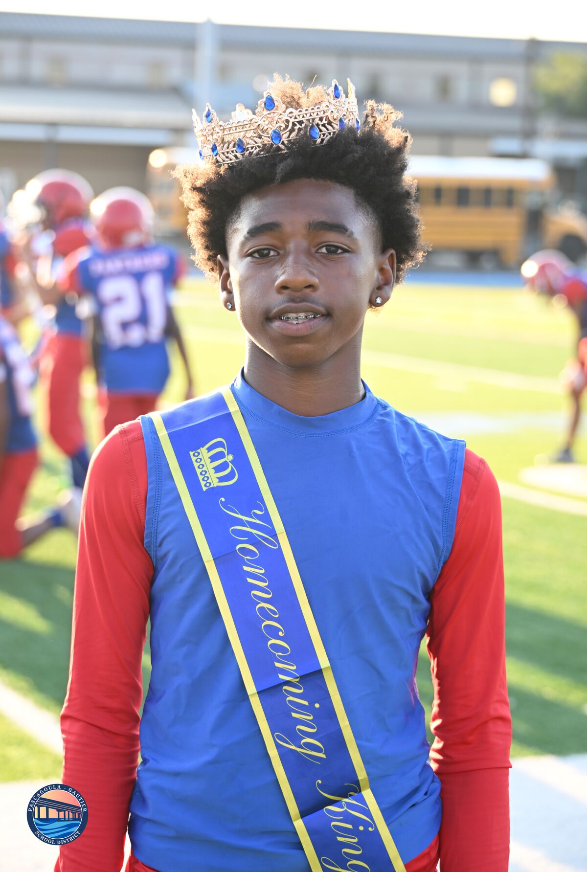 A young man wearing a crown and sash stands on a football field.