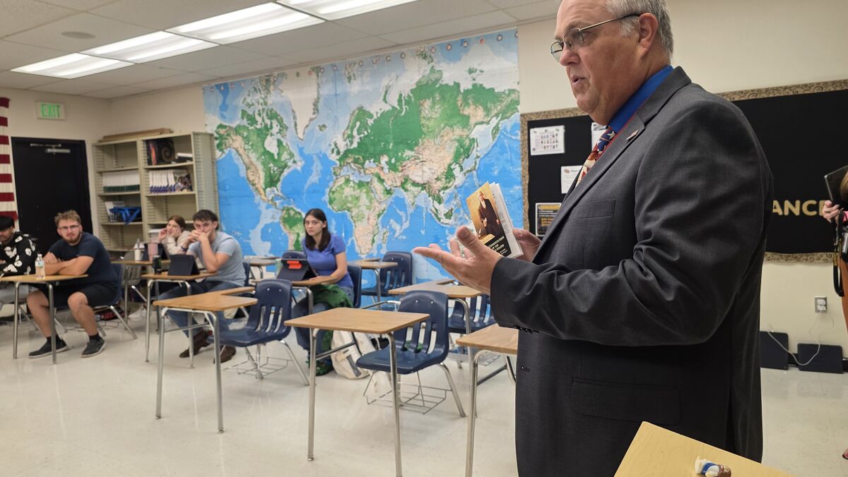 A teacher speaks to students in a classroom, holding a book.