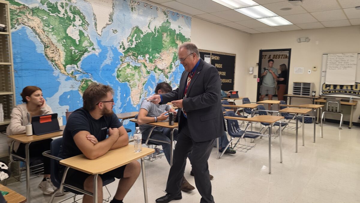 A teacher interacts with students in a classroom, a world map on the wall.