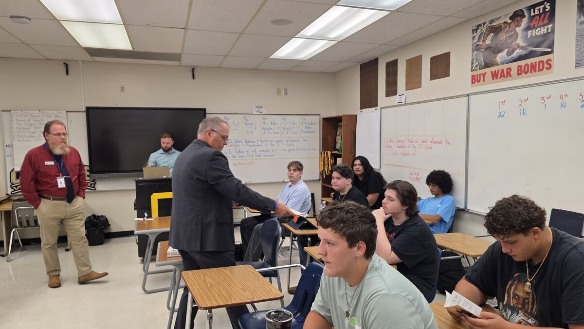 A classroom scene with a teacher addressing students seated at desks.