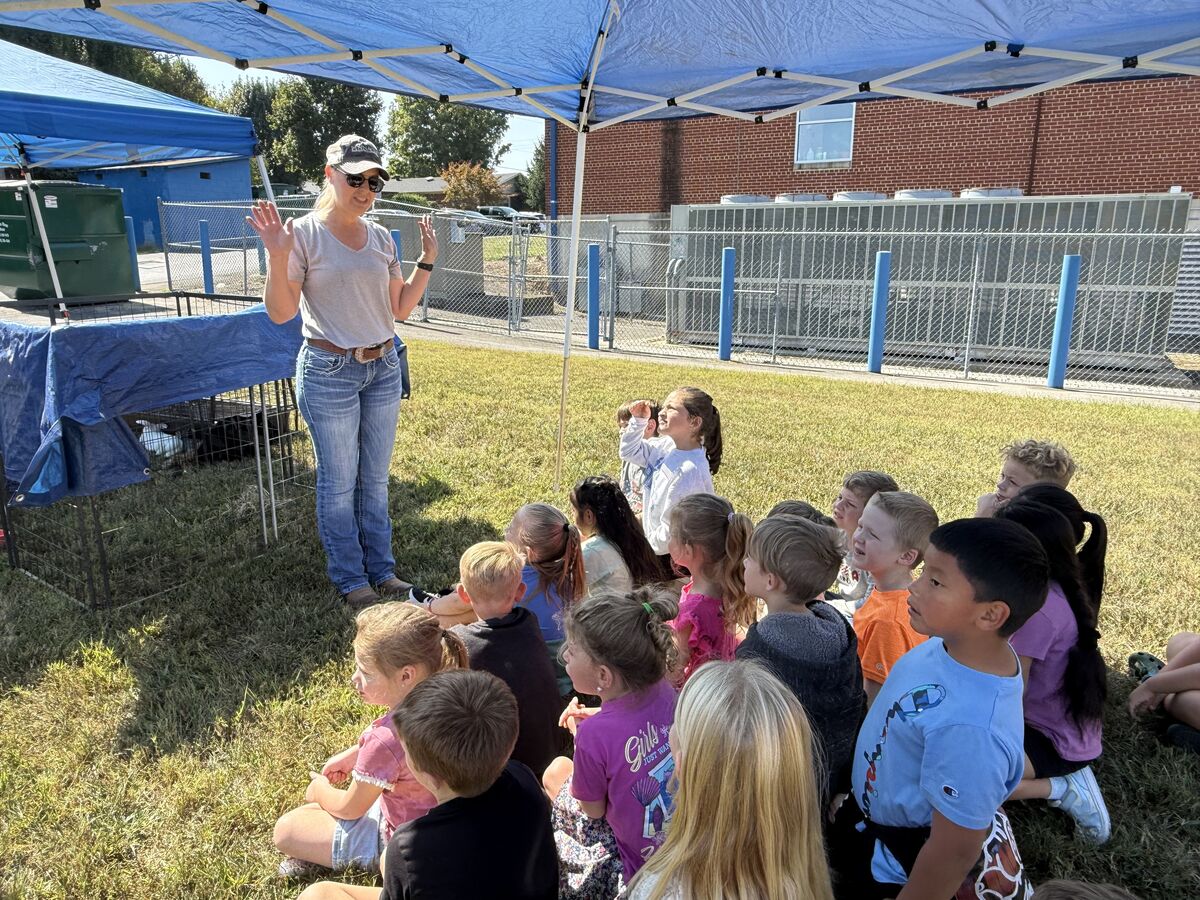A teacher gestures to a group of children seated on the grass under a canopy.