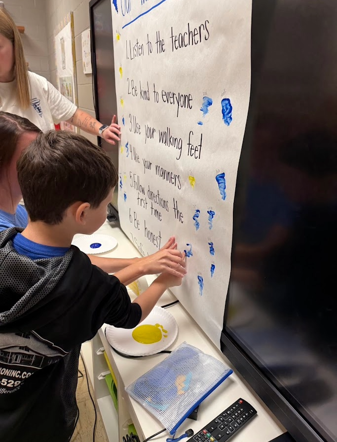 A child adds a blue footprint to a classroom poster about school rules.
