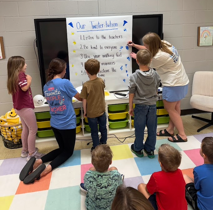 A teacher points to a poster titled 'Our Twister-tution' while students observe.