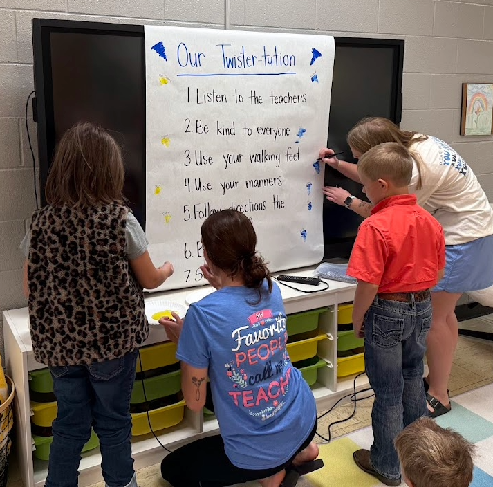 Children and teachers work together on a classroom activity, with a list of rules displayed.
