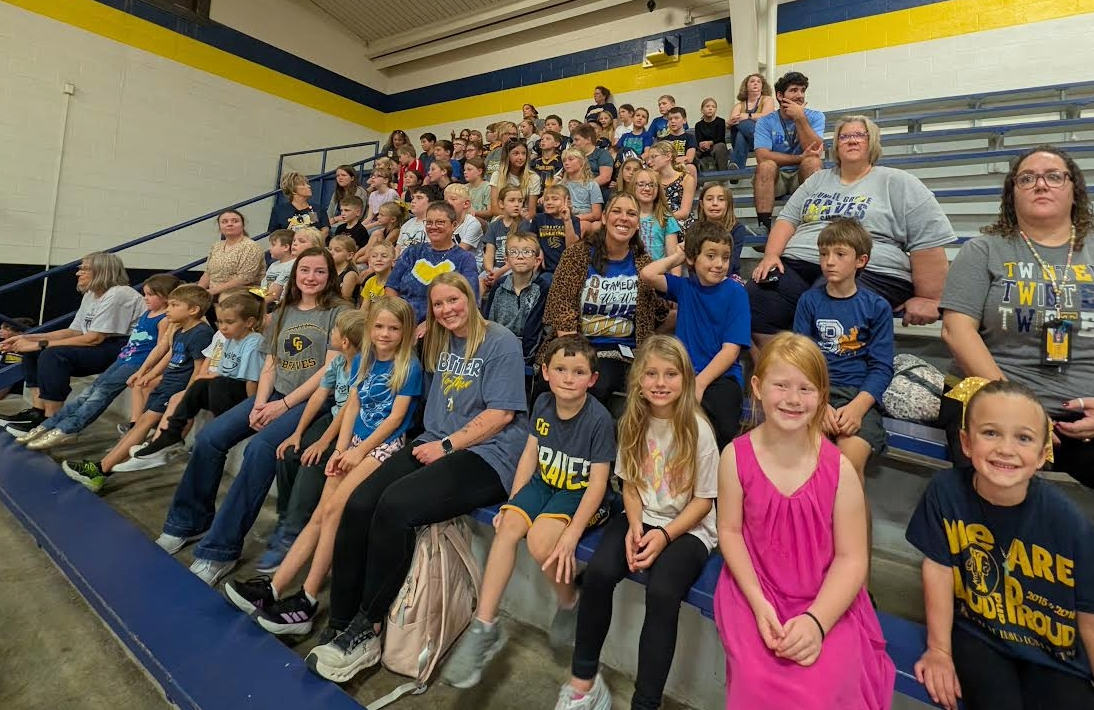A large group of children and adults sit together in bleachers, smiling at the camera.