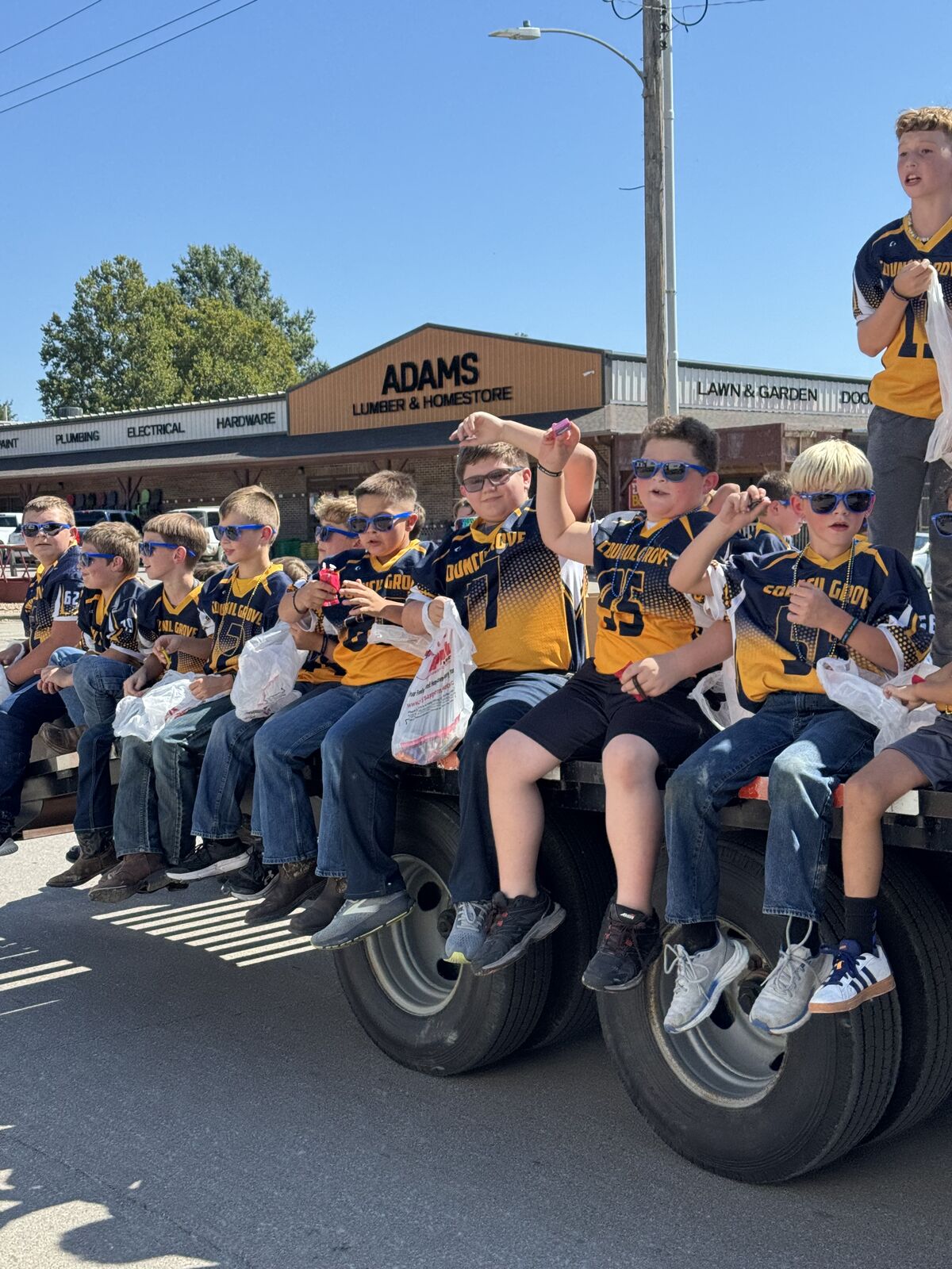 A group of children in matching jerseys ride on a truck bed, wearing sunglasses.