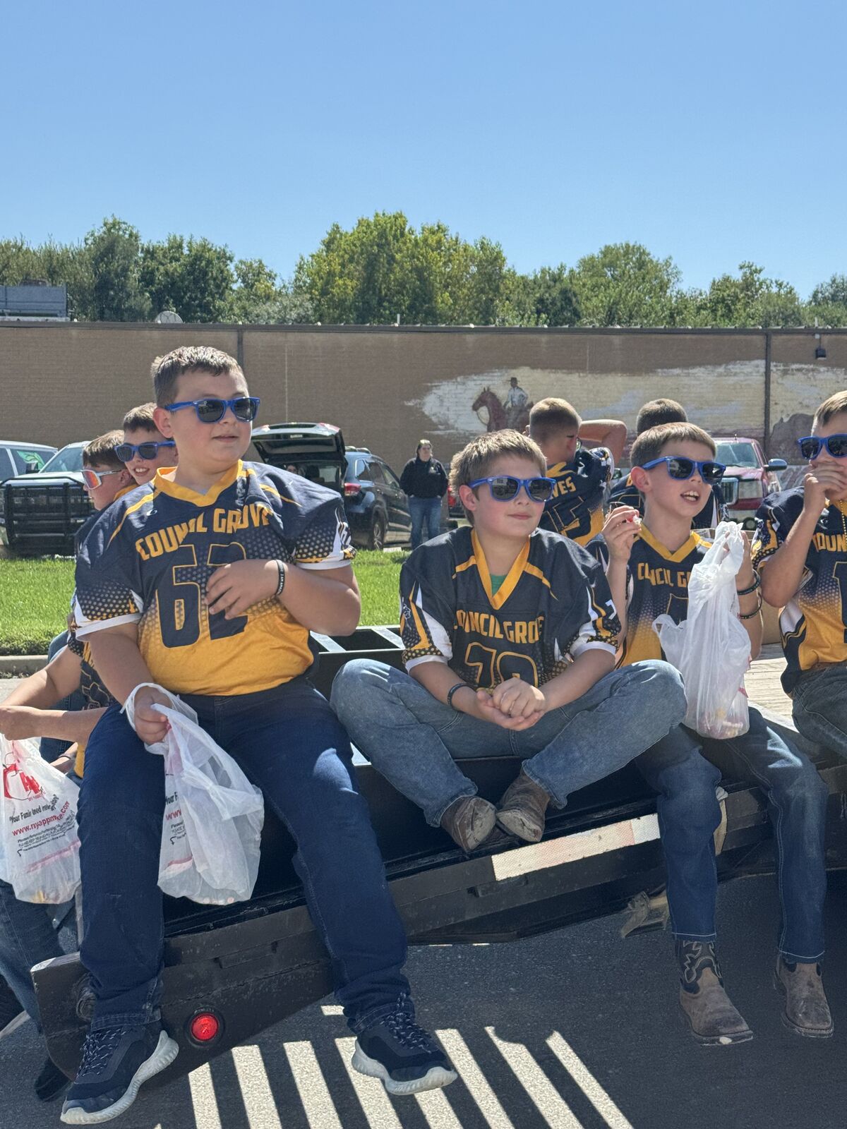 A group of young people wearing sunglasses and jerseys are sitting on a flatbed trailer.