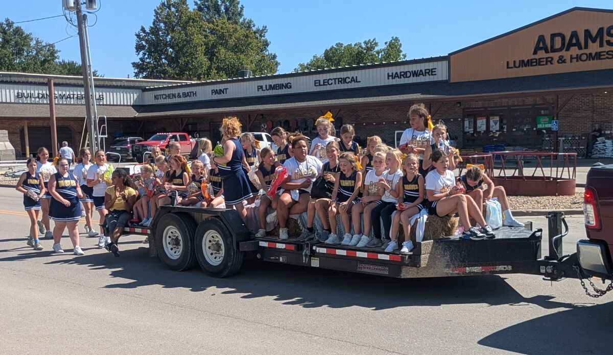 A group of cheerleaders ride on a trailer during a parade, passing by local businesses.