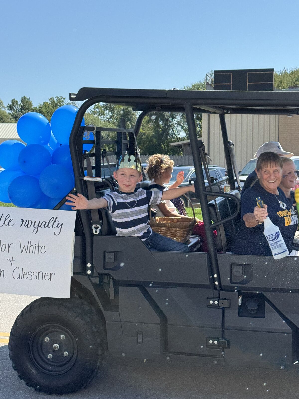 Children and adults ride in a utility vehicle, smiling and waving.