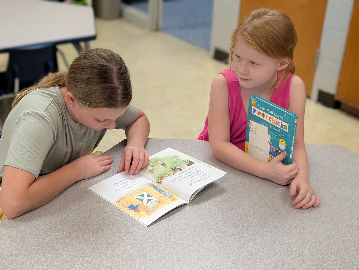 Two young girls read a book together at a table in a classroom.