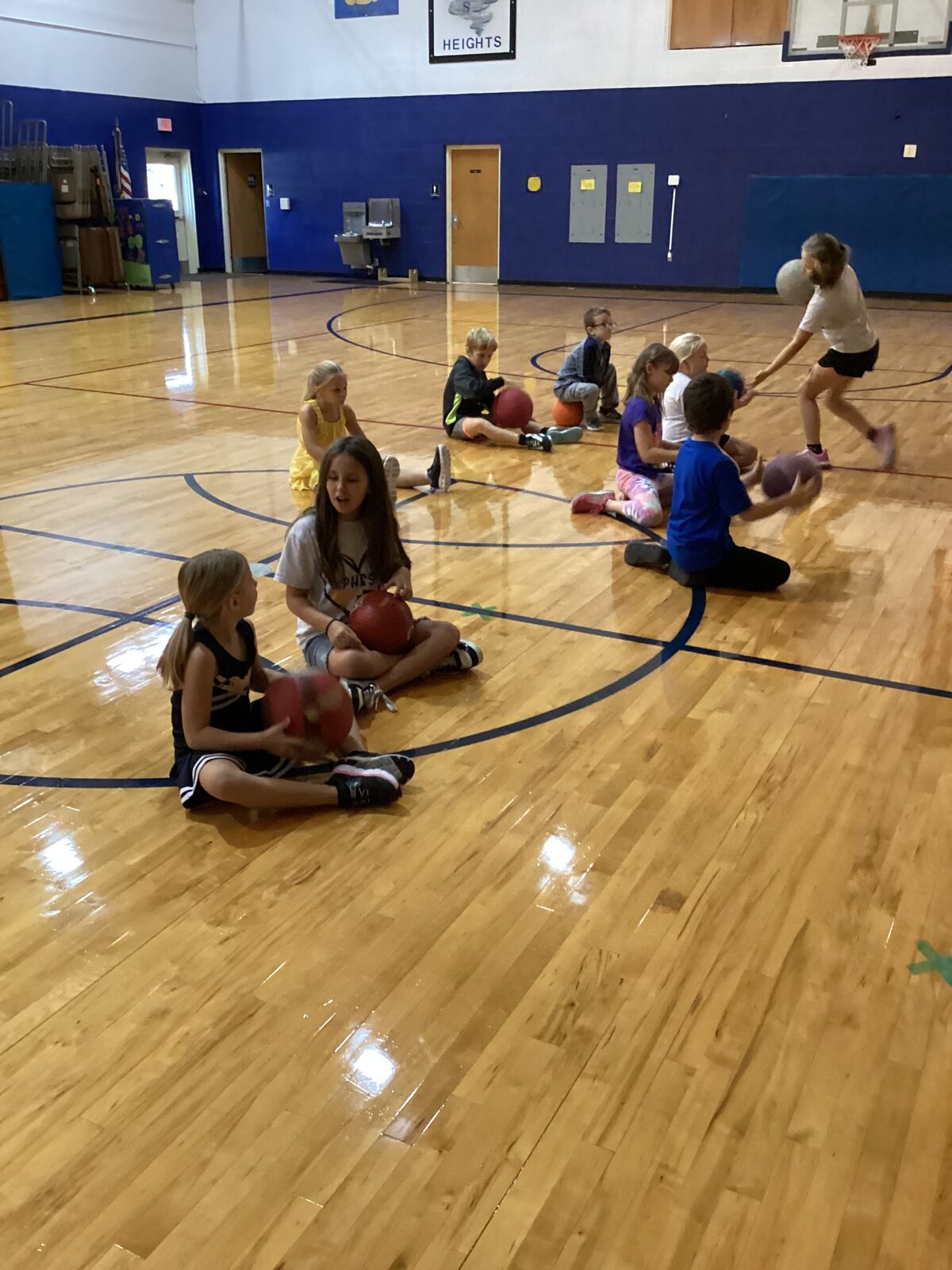 Children sit on a basketball court, some holding red balls.