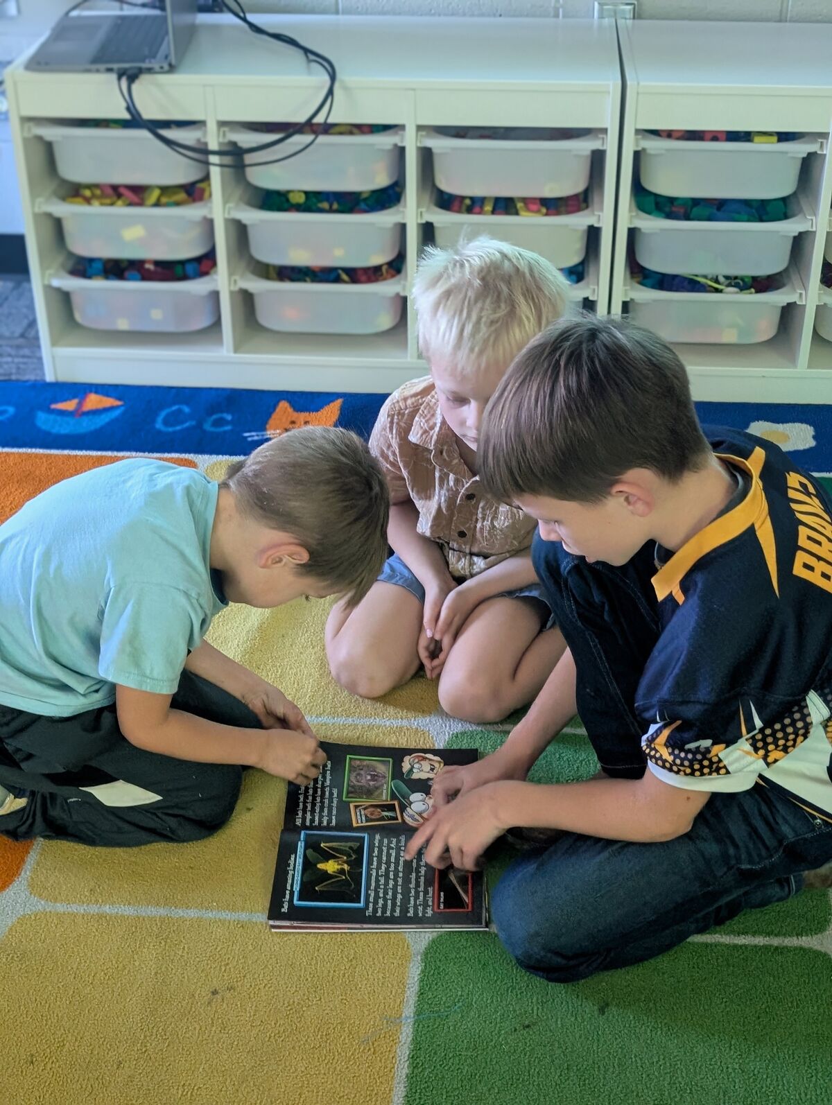 Three children sit on a colorful rug, looking at a book together.