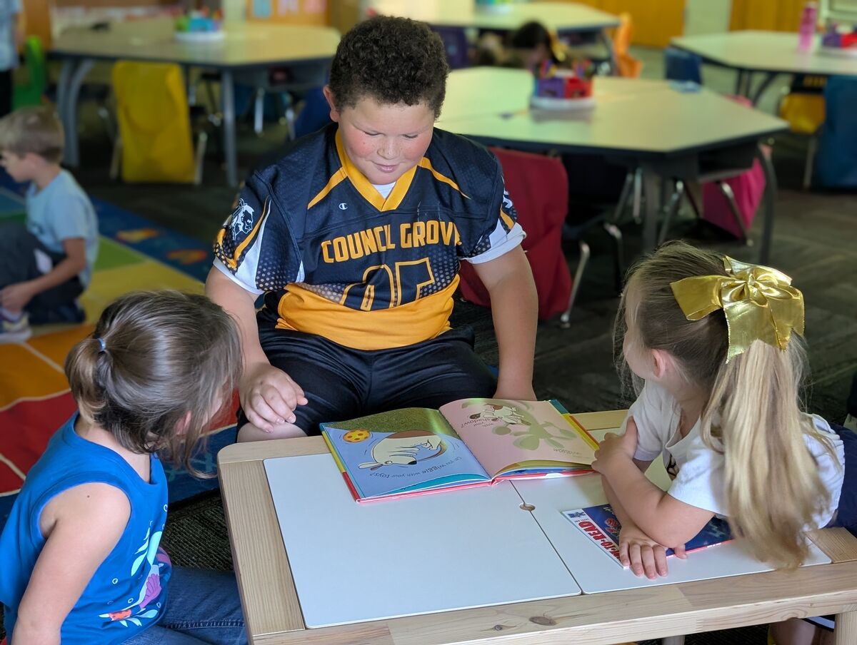 Children gather around a table, looking at a book together in a classroom.