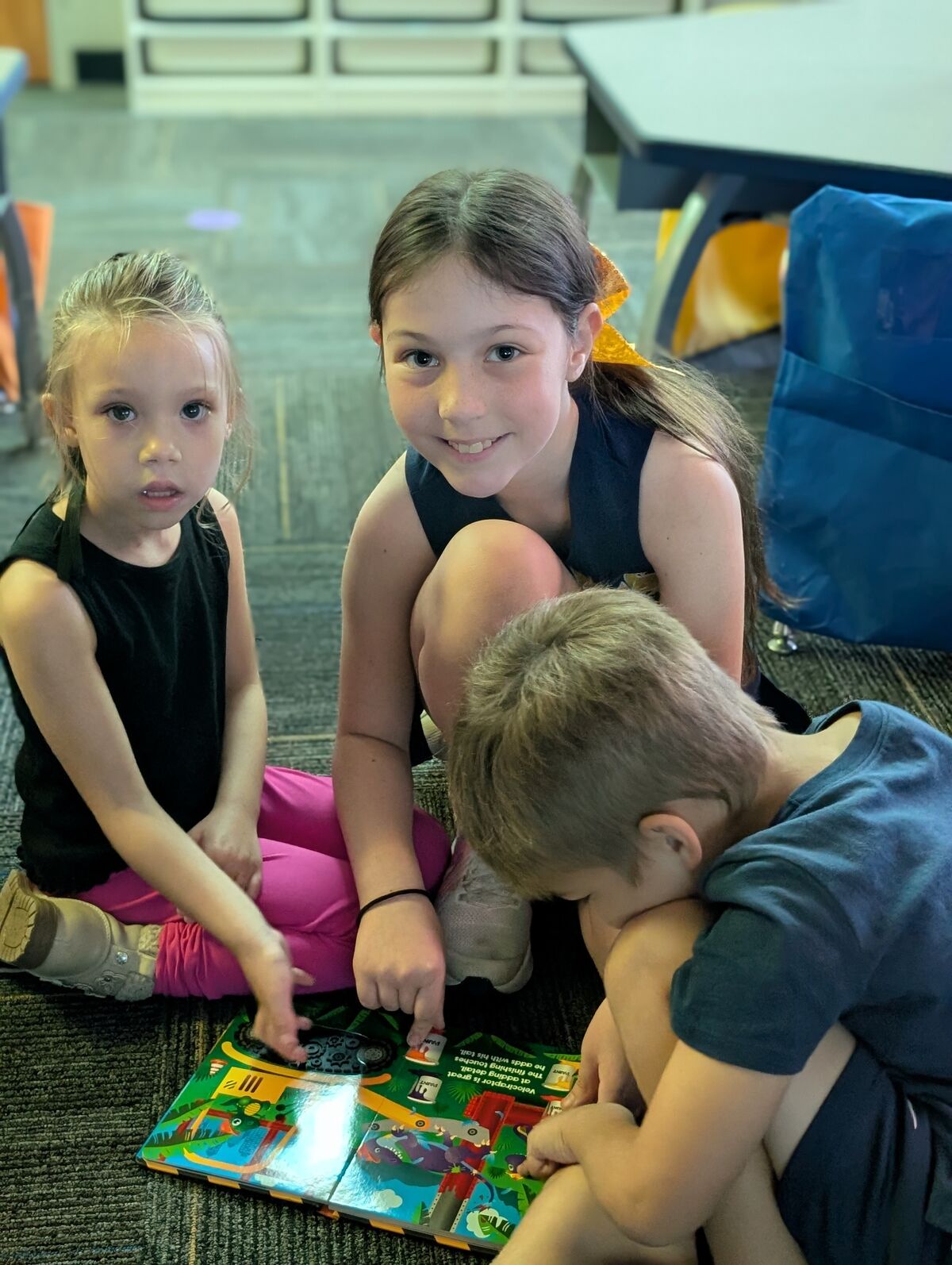 Three children sit on the floor, looking at a colorful book together.