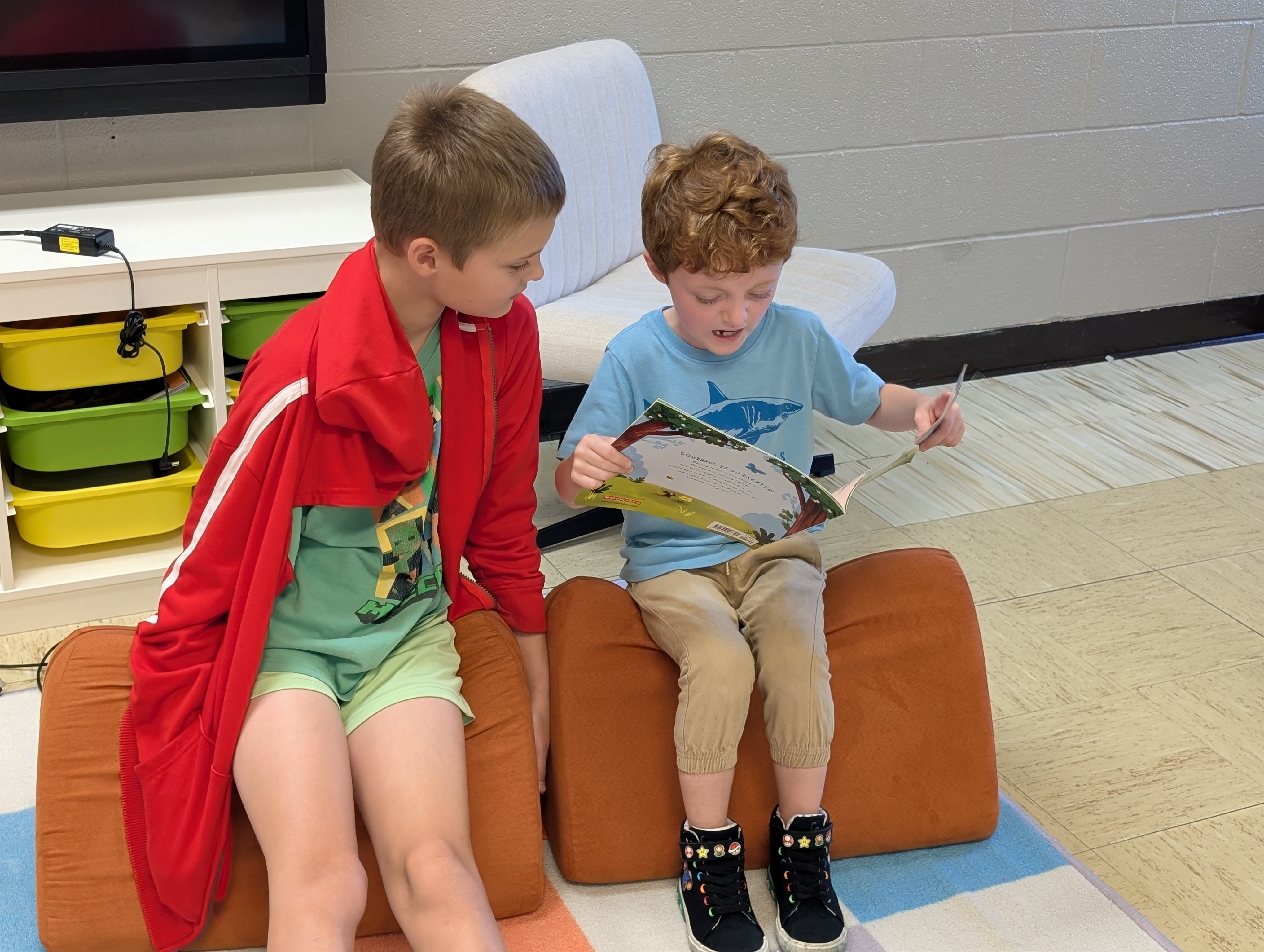 Two young boys sit on cushions, one reading a book with excitement.