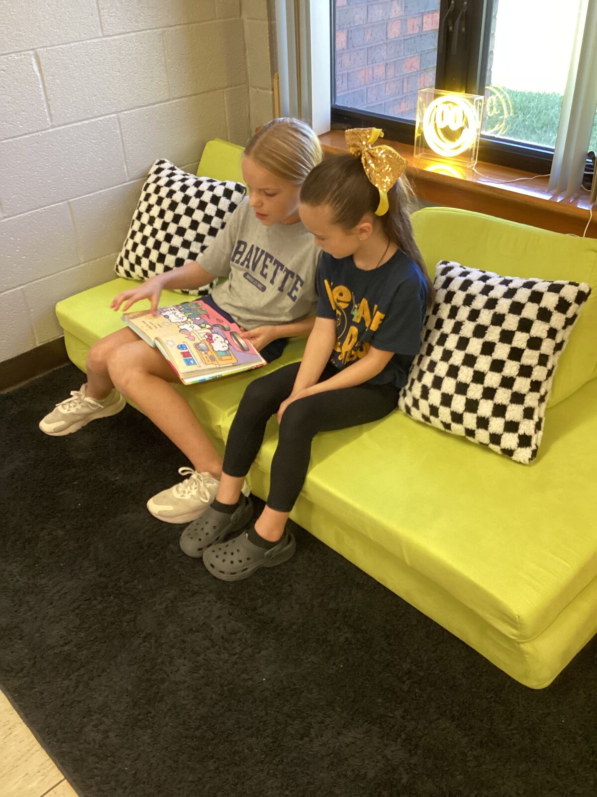 Two young girls read a book together while relaxing on a lime green couch.