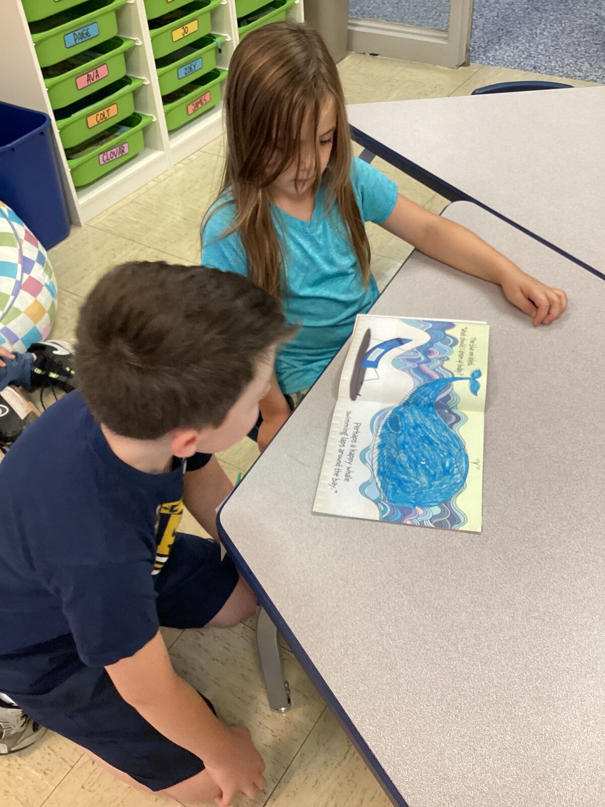 Two children read a book together at a desk, focusing on a whale illustration.