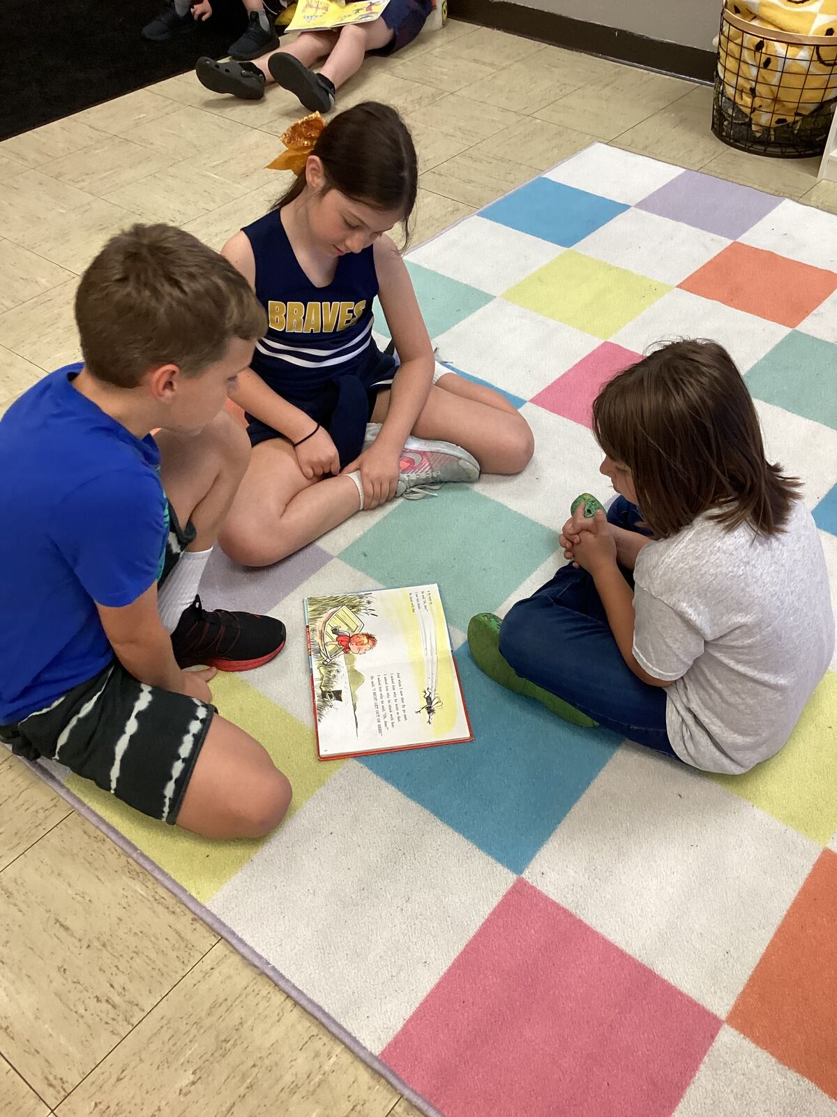 Three children sit on a colorful rug, reading a book together.