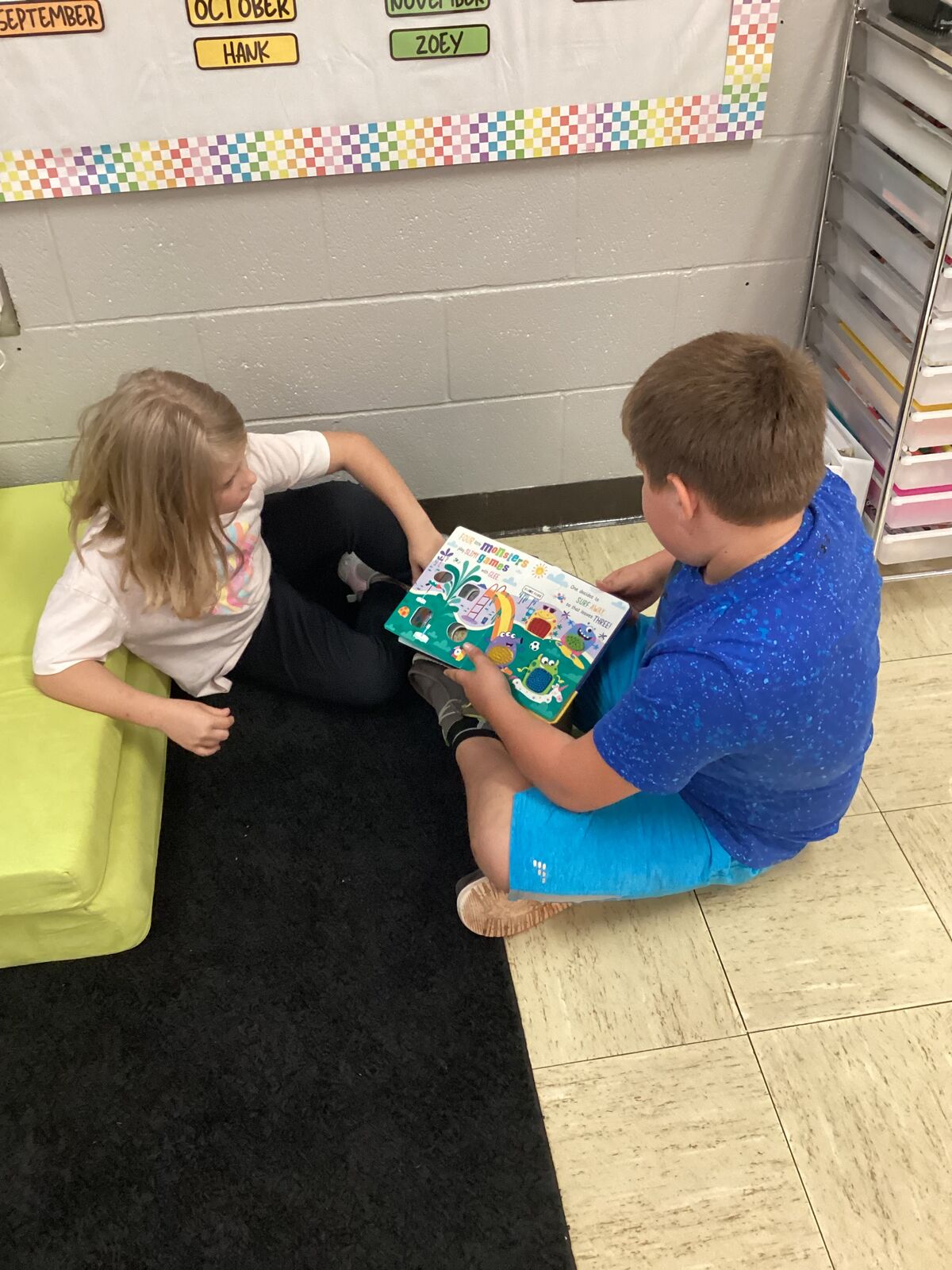 A boy and girl read a colorful book together in a classroom setting.