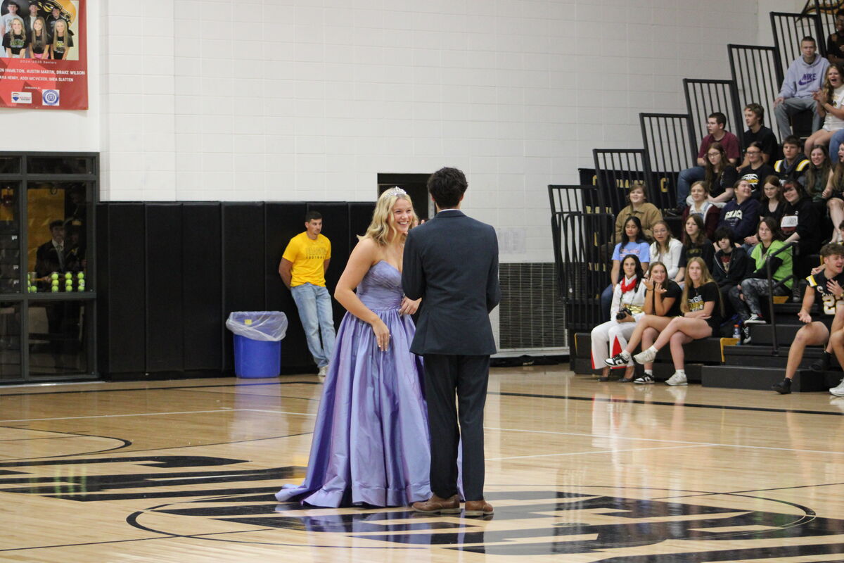 Senior attendant in a formal gown smiles at a young man in a suit on a basketball court.