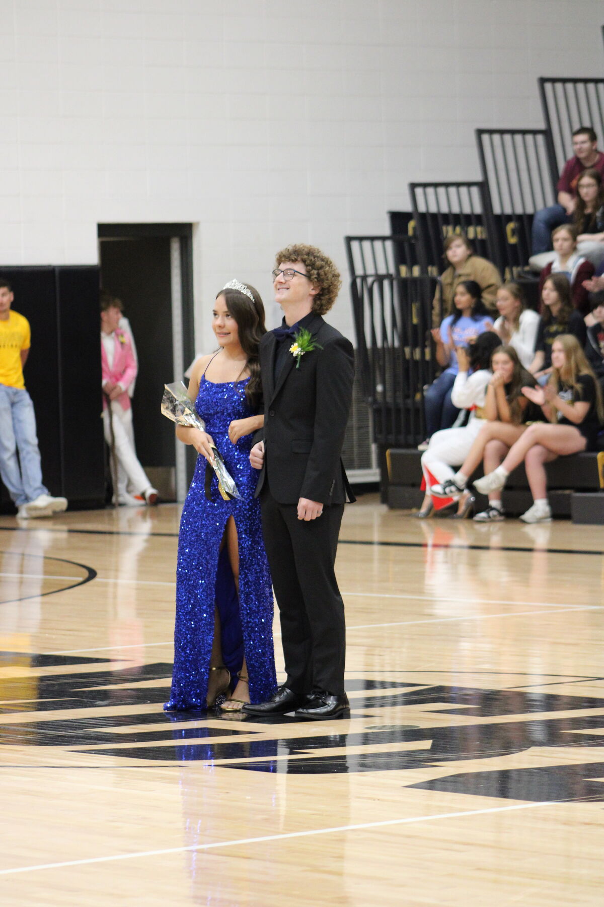 Senior king and queen candidate stand on a basketball court, the woman in a blue gown, the man in a suit.