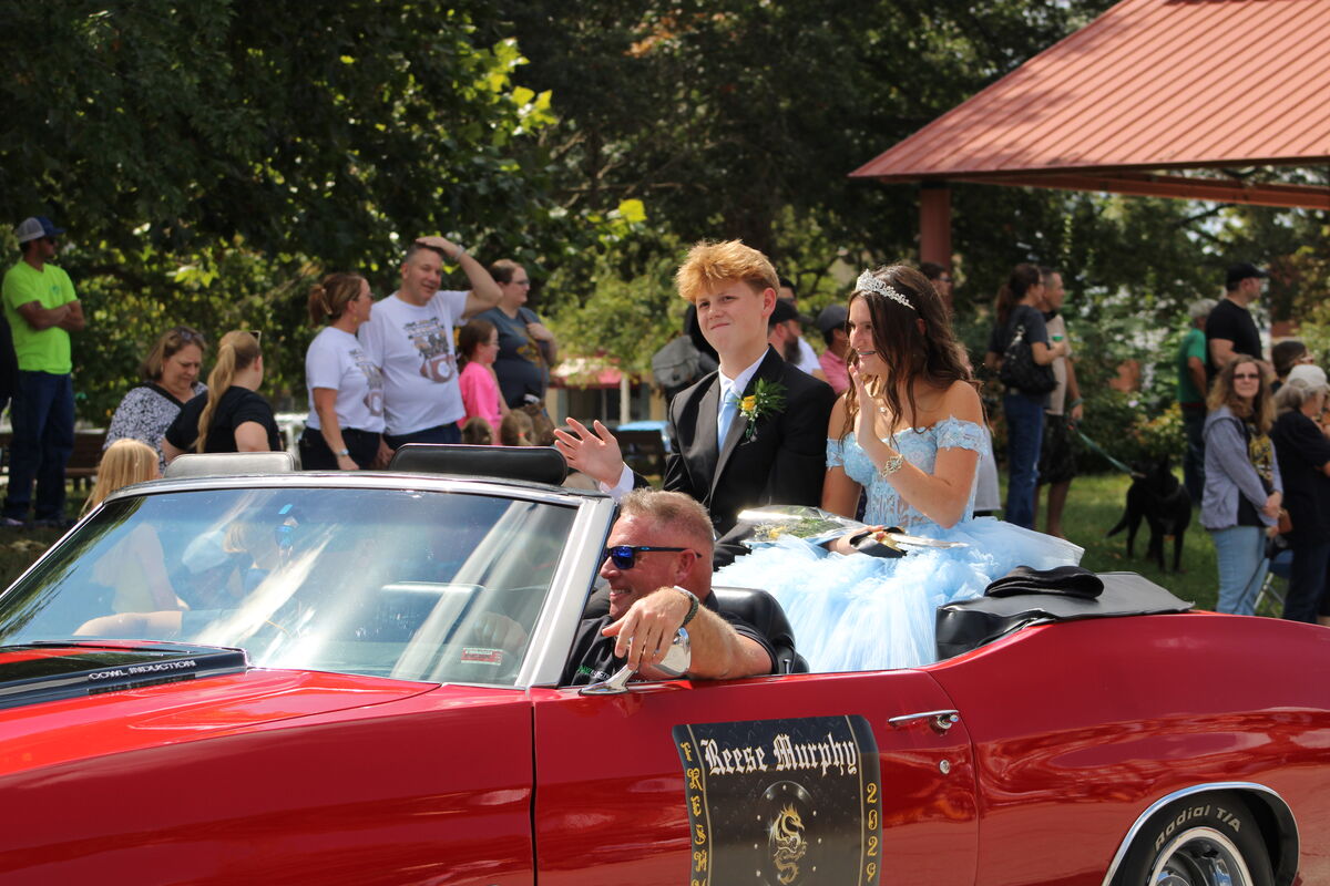 Freshmen royalty dressed in formal attire ride in a classic red convertible, waving to onlookers.