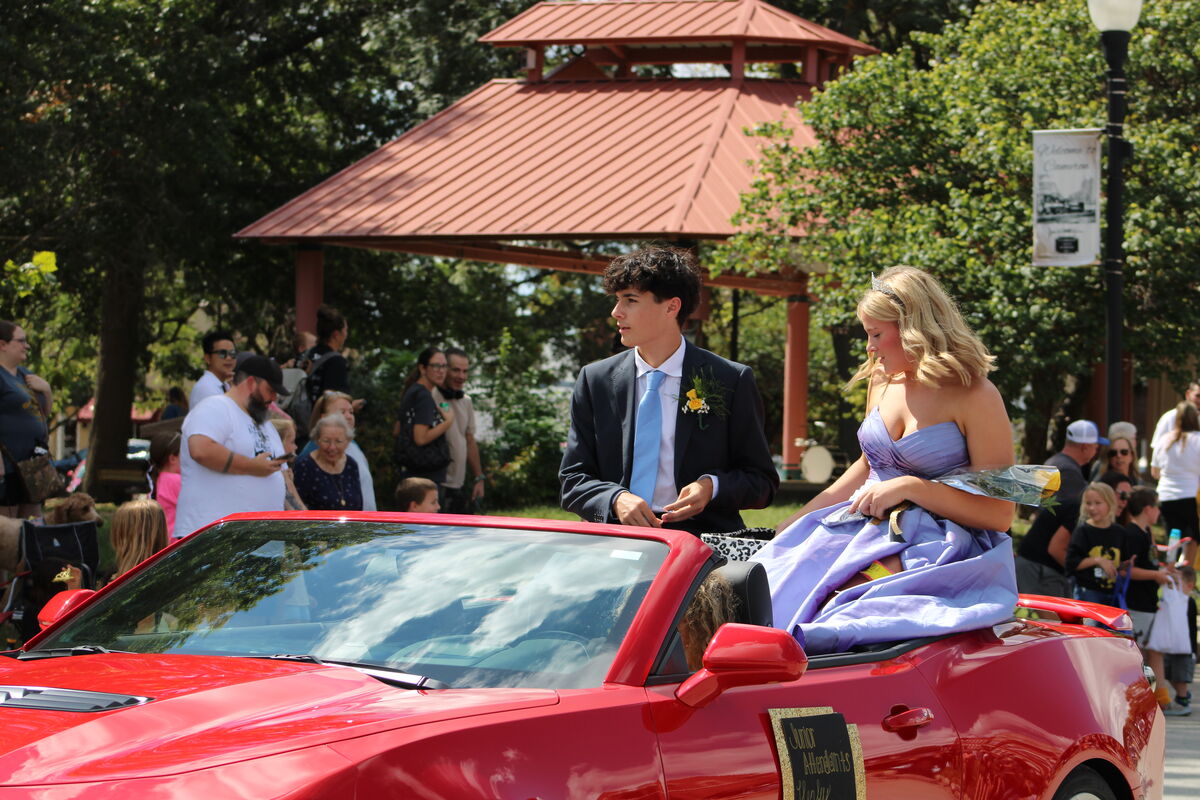 A young couple in formal attire ride in a red convertible during the homecoming parade