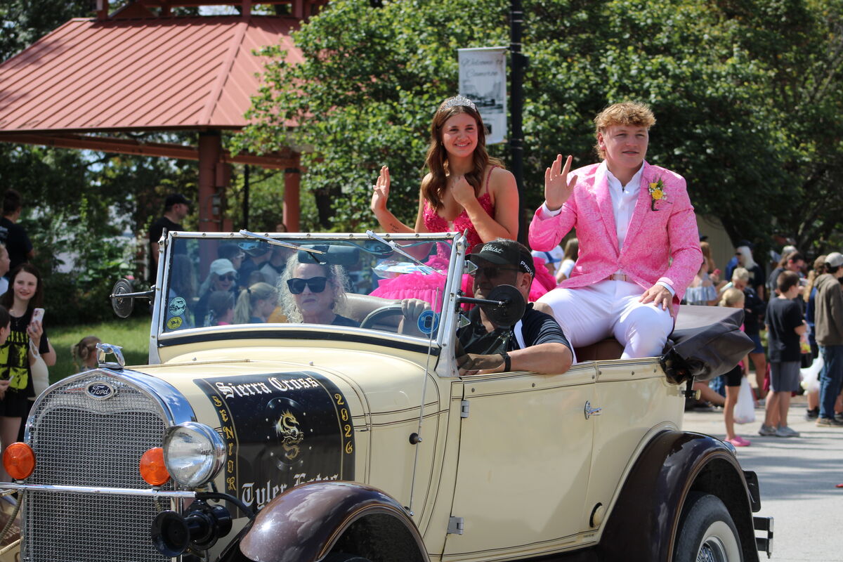 A classic car carries a young woman in a pink dress and a young man in a pink suit, waving to a crowd.