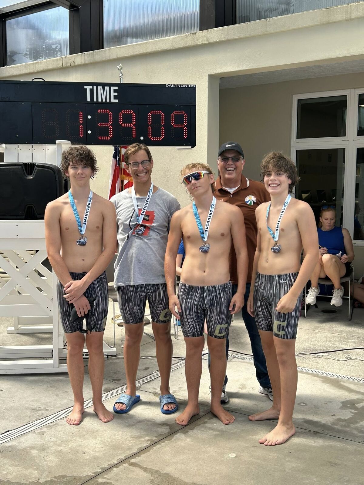 A group of swimmers pose with medals after a competition, the scoreboard reads 1:39.09.