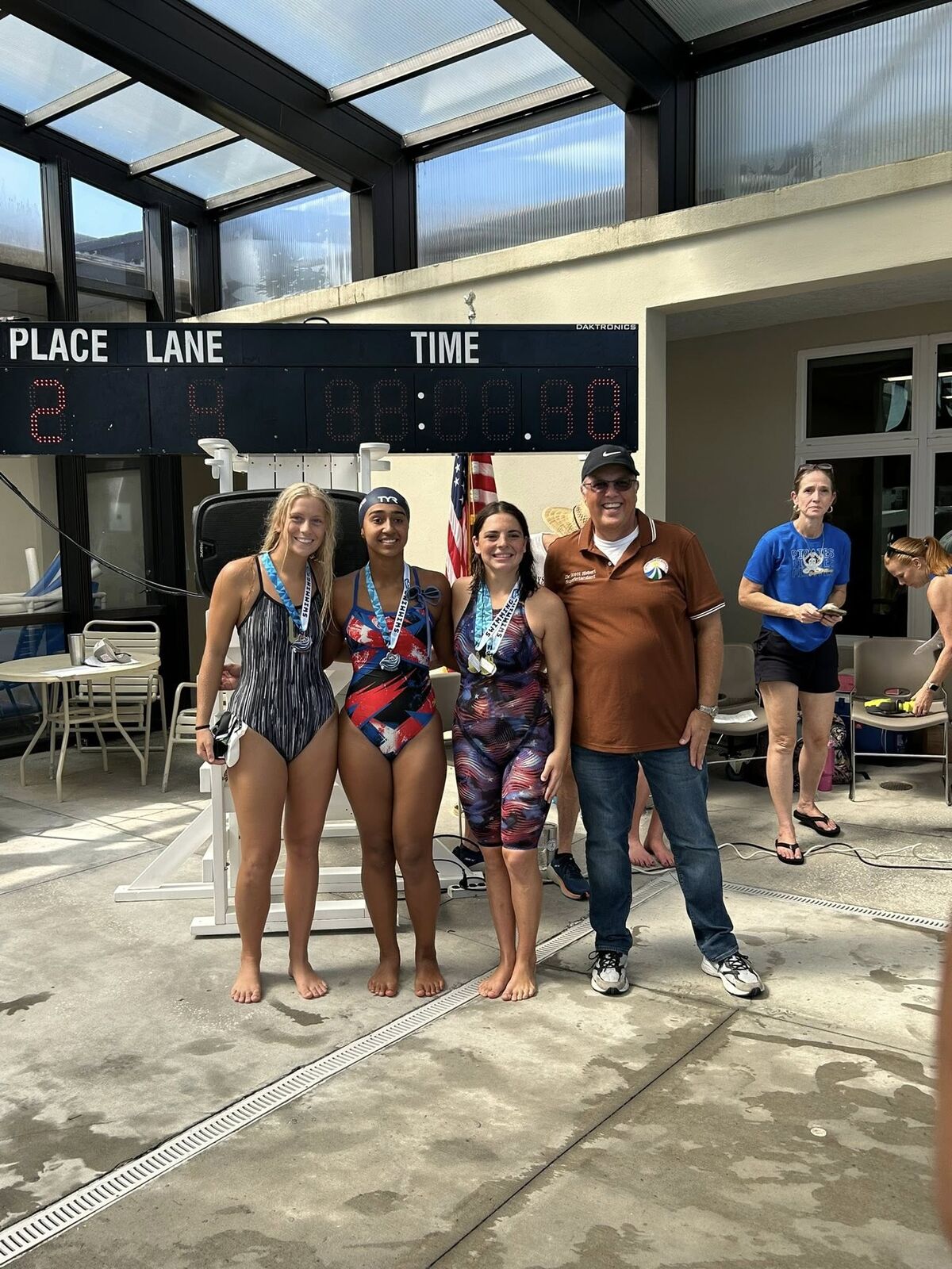 Three swimmers pose with a man, all wearing medals, near a scoreboard.