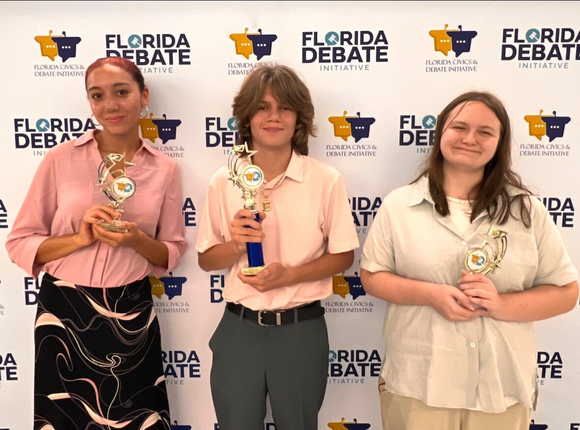 Three young people stand proudly, each holding a trophy, in front of a 'Florida Debate Initiative' backdrop.