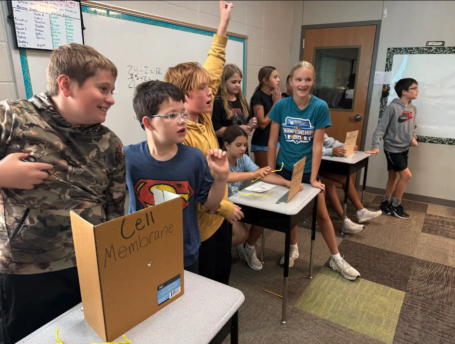Students in a classroom participate in a science activity, some raising their hands.