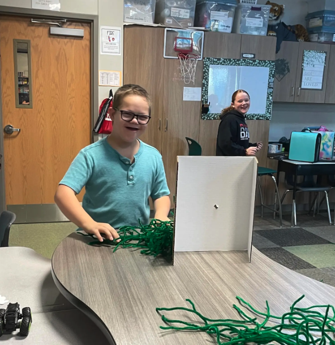 A student smiles while working on a craft project in a classroom.