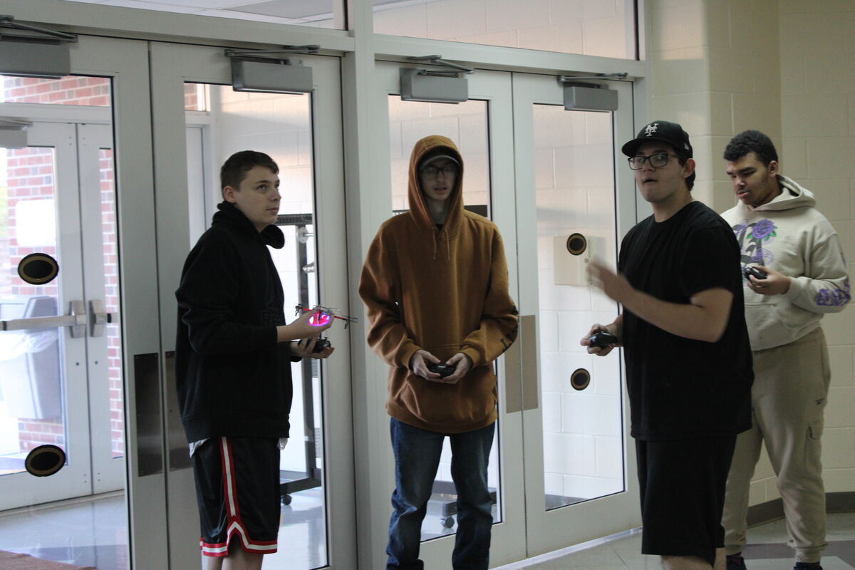 Four young people stand near glass doors, holding controllers, possibly for a drone.