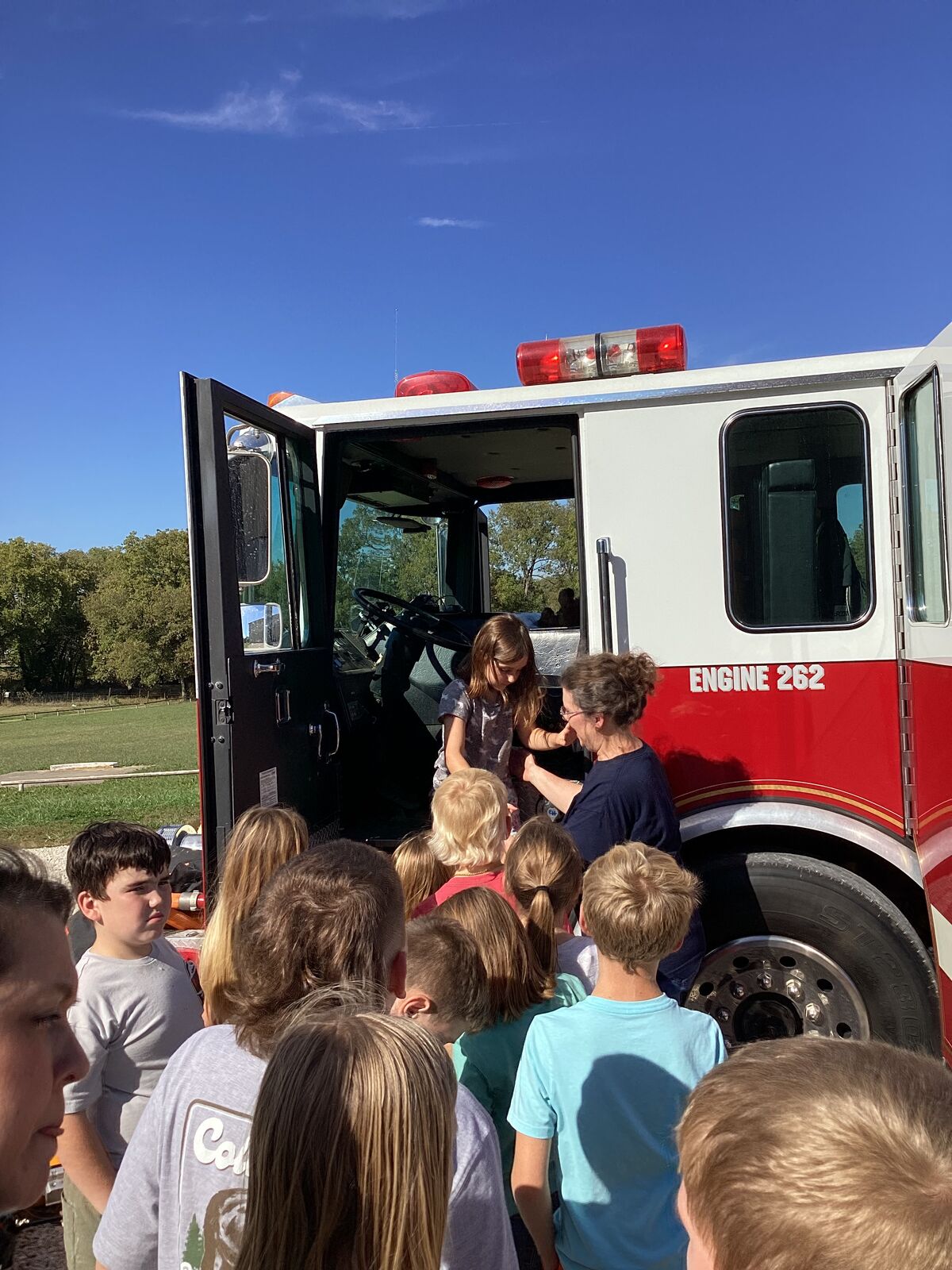 Children gather around a fire truck with an open door, under a clear blue sky.