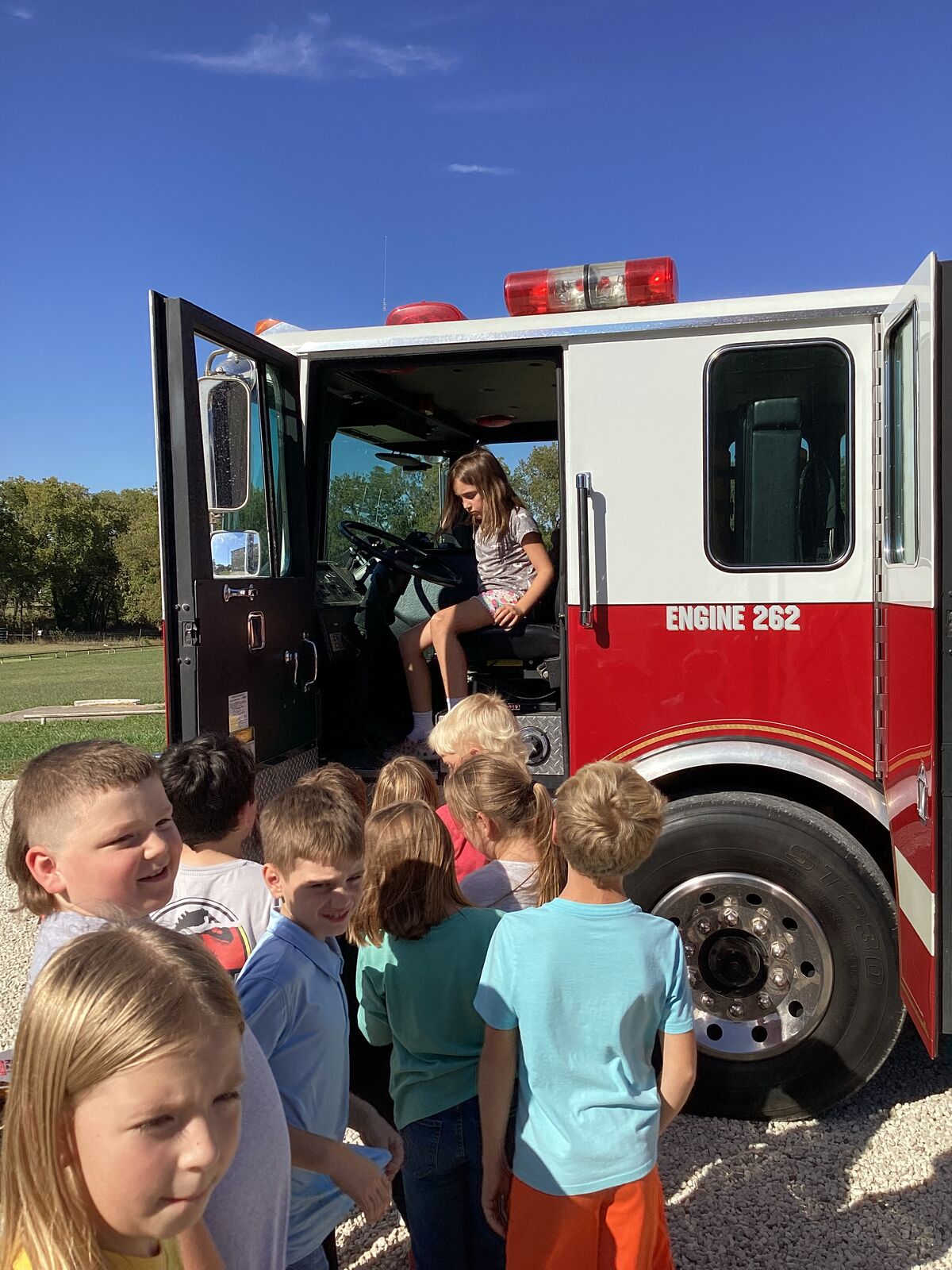 Children gather around a fire truck, with one child sitting in the driver's seat.