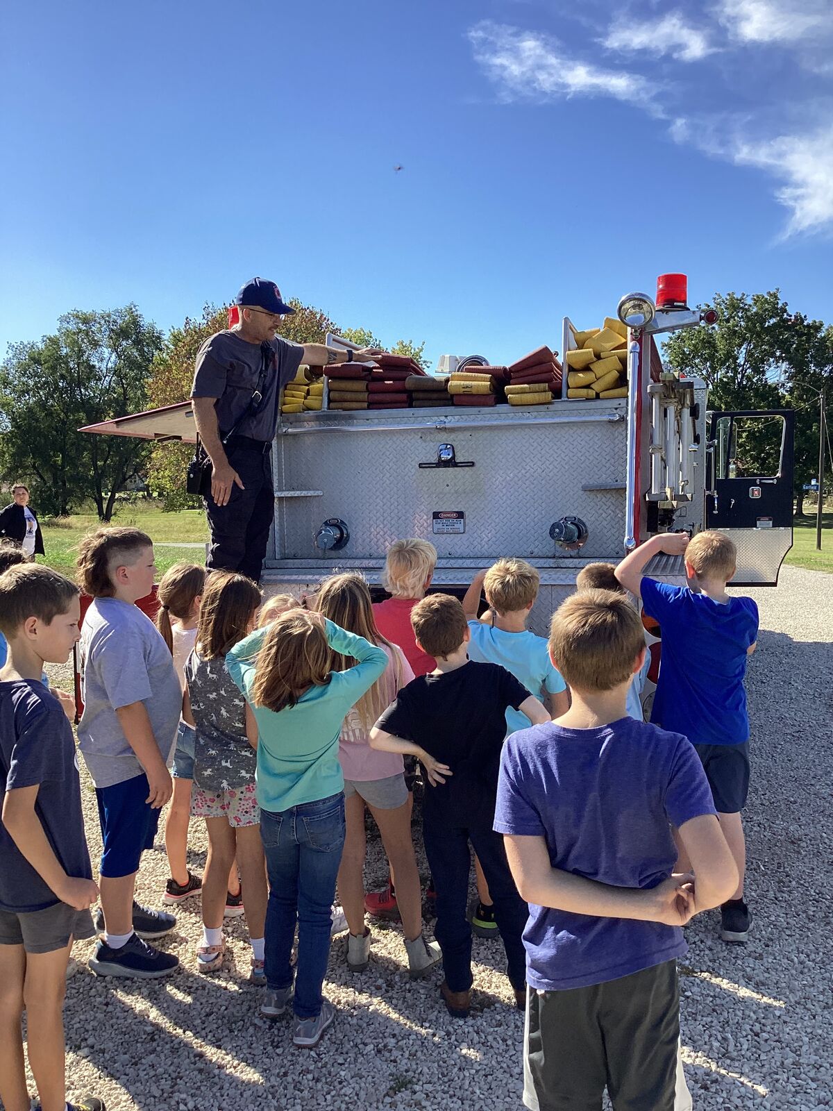 A firefighter points to equipment on a fire truck while children watch attentively.