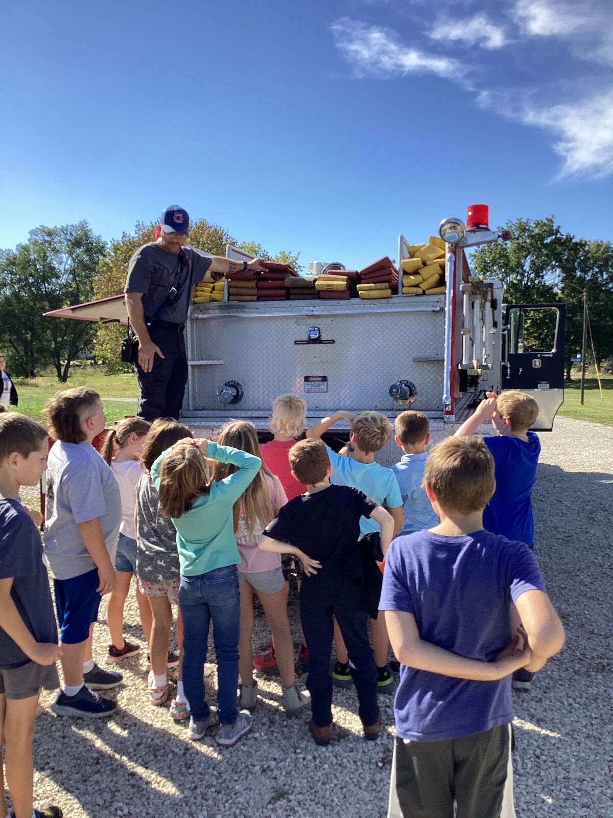 A firefighter shows a group of children the equipment on a fire truck on a sunny day.