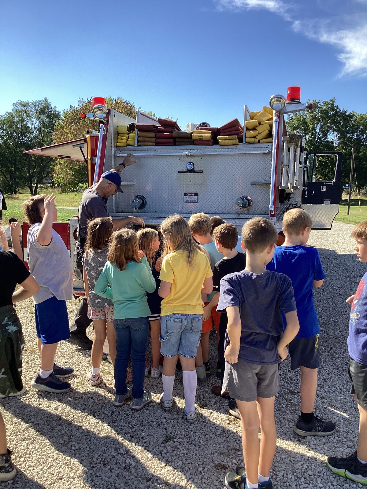 A group of children gather around a fire truck, listening to an adult.