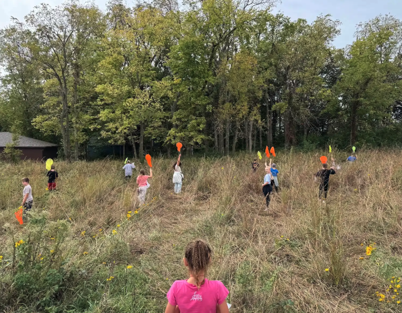 Children explore a grassy field, holding up colorful flags near a wooded area.