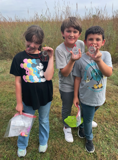 Three children hold up compasses in a grassy field, smiling at the camera.