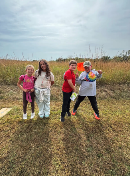Four children pose for a photo in a grassy field on a cloudy day.