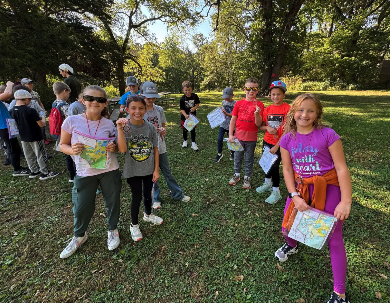 A group of children smile while holding maps and compasses outdoors.