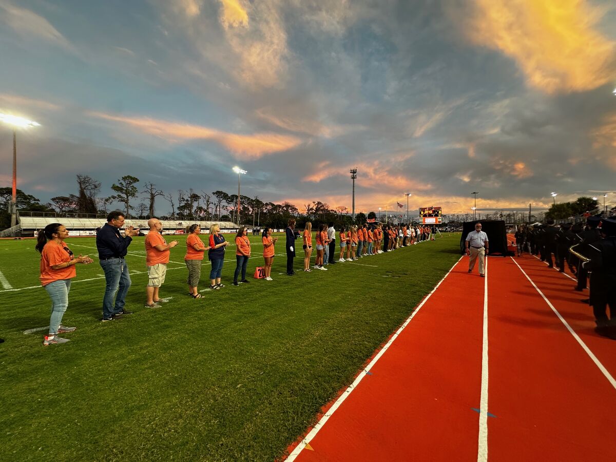 People stand on a football field, possibly at a graduation, under a colorful sunset.